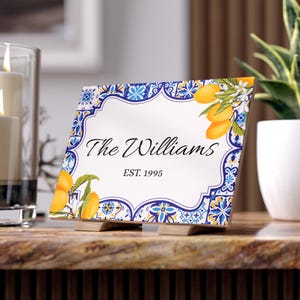 May include: A decorative ceramic tile with a white centre, blue and yellow border, and lemon illustrations. The tile reads "The Williams" and "EST. 1995" in black script. It is propped up on two small wooden stands.