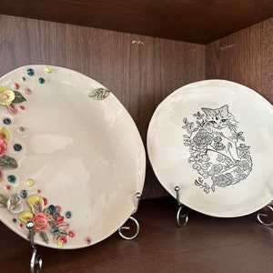 Puede incluir: Dos platos de cerámica blanca con diseños decorativos. Un plato presenta un diseño floral con flores y hojas en relieve en tonos rojos, amarillos y azules. El otro plato presenta una ilustración en blanco y negro de un gato sentado en un arreglo floral.