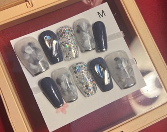 May include: A set of ten press-on nails in a clear plastic case. The nails are in a variety of styles, including glitter, marble, and solid colors. The case has a label that says "M".