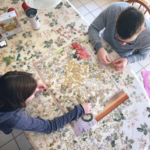 May include: Two people working on a large jigsaw puzzle with a floral pattern on a white tablecloth. The puzzle is almost complete and the pieces are scattered around the table.