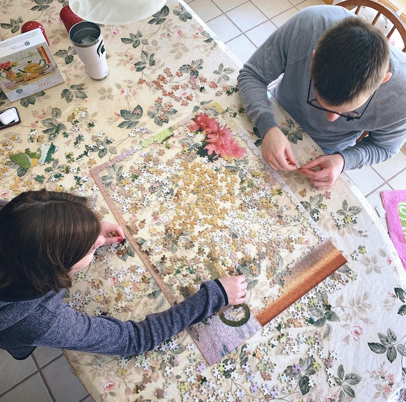 May include: Two people working on a large jigsaw puzzle with a floral pattern on a white tablecloth. The puzzle is almost complete and the pieces are scattered around the table.