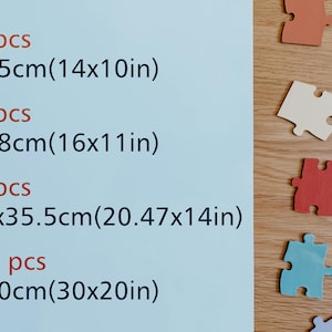 May include: A blue background with text listing the number of pieces and dimensions of jigsaw puzzles in inches and centimeters. The text reads: 200 pcs 35x25cm(14x10in) 300 pcs 40x28cm(16x11in) 500 pcs 52.5x35.5cm(20.47x14in) 1000 pcs 75x50cm(30x20in). The image also shows a wooden surface with several colorful jigsaw puzzle pieces.