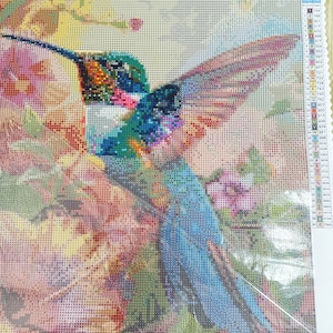 May include: A colourful diamond painting kit featuring a hummingbird with a long beak and iridescent blue, green, and red feathers. The hummingbird is perched on a branch with pink flowers. The kit includes a colour chart with corresponding numbers for each diamond.