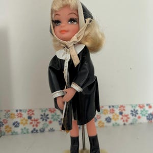 May include: A blonde doll with painted facial features, long eyelashes, and orange lips. The doll wears a black shiny coat with a white collar, a matching black headscarf tied under the chin, and black boots. It stands on a circular base.