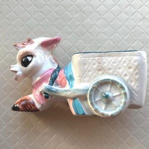 May include: A ceramic planter shaped like a donkey pulling a cart. The donkey is white with pink ears, a pink chest, and brown hooves. The cart is white with a textured diamond pattern on the side and features a single light blue wheel on each side.
