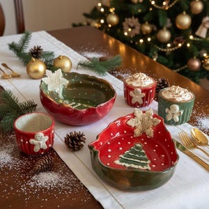 Può includere: Un'impostazione festiva della tavola di Natale con ciotole e tazze in ceramica rosse e verdi. Le ciotole presentano disegni di fiocchi di neve e alberi di Natale. Le tazze sono decorate con omini di pan di zenzero. Posate e ornamenti dorati completano la scena.