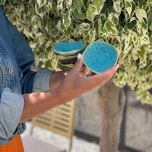 Op de afbeelding: Een stapel kleine, handgemaakte keramische kommen. De bovenste kom is helder turkoois met een textuur en een gouden rand. De kommen eronder hebben een donkerbruin en groen kleurenschema. De kommen worden in handen gehouden.