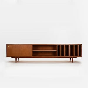 May include: A mid-century modern wooden media console in a warm brown hue. It has a sliding door, open shelving, and vertical dividers for storage. The console is supported by tapered legs and a brass-coloured knob.