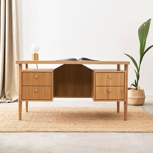 Peut inclure: Un bureau en bois clair avec deux ensembles de tiroirs, chacun avec deux tiroirs. Le bureau a un design minimaliste et est placé sur un tapis tissé. Une lampe et une plante sont également visibles.