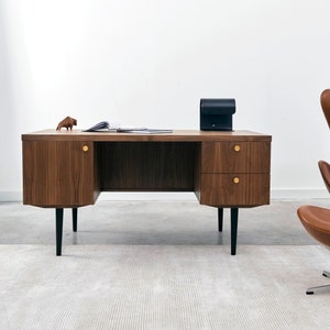 Könnte beinhalten: Ein Schreibtisch aus Walnussholz im Mid-Century-Modern-Stil mit zwei Schubladen und zwei Schränken, Messingbeschlägen und schwarzen Beinen. Ein brauner Ledersessel und eine kleine Holzfigur eines Bisons sind ebenfalls zu sehen. Der Schreibtisch steht auf einem hellen Teppich.