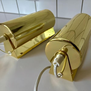 May include: Two gold wall sconces with cylindrical shades and rectangular bases. Each sconce has a white electrical cord and a gold metal frame. The sconces are designed to be mounted on a wall.