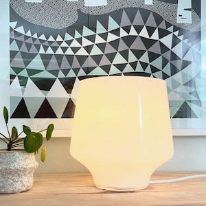 May include: A white, opaque table lamp with a geometric, faceted base. The lamp is turned on and illuminating the room. The lamp is sitting on a wooden surface next to a potted plant.