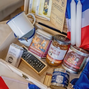 May include: A wooden box filled with vintage items, including enamel mugs, rationed chocolate, tea, and hot chocolate tins, a jar of orange jam, dominoes, matches, candles, and a flag. The items are arranged inside the box.