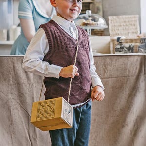 May include: A child in a brown patterned knitted vest, white shirt, and blue shorts, wearing a newsboy cap. They are holding a small cardboard box with text and illustrations, suspended by a rope. The background suggests a vintage shop or cafe setting.