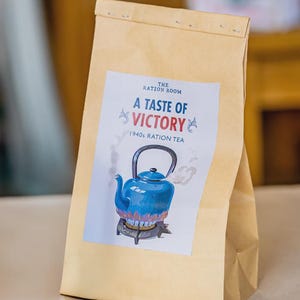 Può includere: Un sacchetto di carta marrone con un'etichetta bianca con la scritta "A Taste of Victory" e "1940s Ration Tea". L'etichetta presenta un'illustrazione di una teiera blu con vapore. Il sacchetto è sigillato in alto.