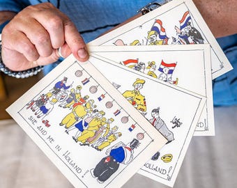 May include: A stack of vintage-style postcards featuring cartoon illustrations of people and Dutch flags. The postcards have the text "SHE AND ME IN HOLLAND!" and other Dutch-themed imagery. The cards are held by a person wearing a blue shirt.