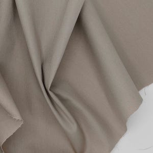 May include: A piece of solid taupe-colored fabric with a visible woven texture. The material is draped in soft, flowing folds, creating natural shadows and highlights across the surface. The edges of the textile show a slightly frayed appearance.