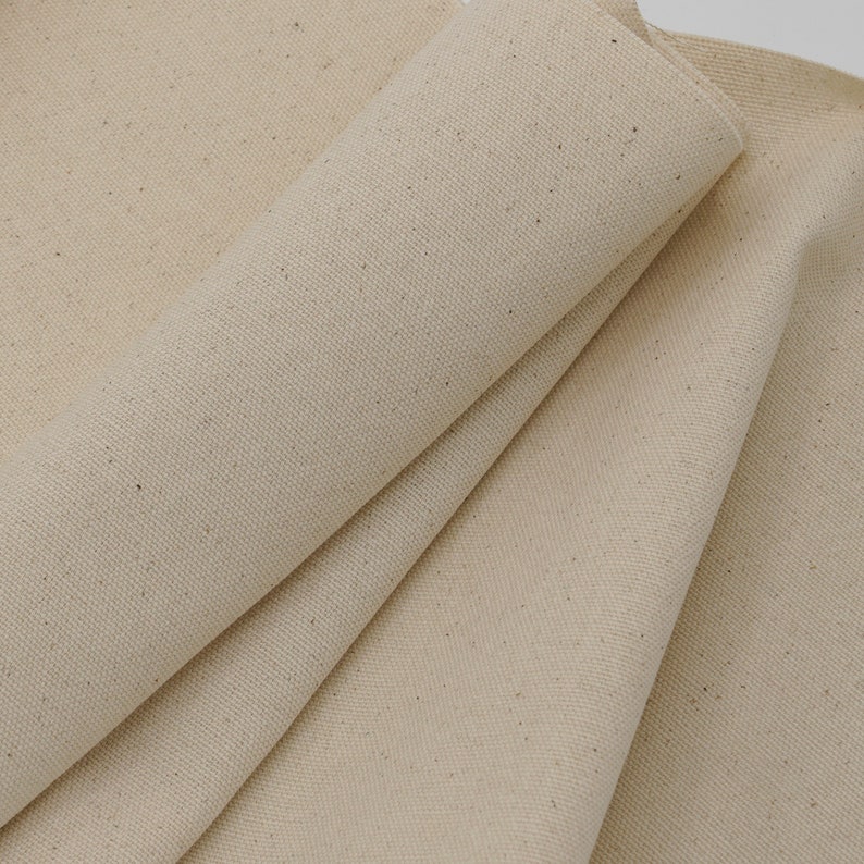 May include: Close-up of a natural, off-white canvas fabric with a subtle texture. The fabric is folded and shows the weave of the material.