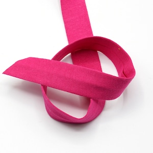 May include: A length of vibrant fuchsia pink bias tape, looped and arranged against a white background. The fabric appears to be a soft, knit material. The tape is folded over on itself, creating a rounded shape.