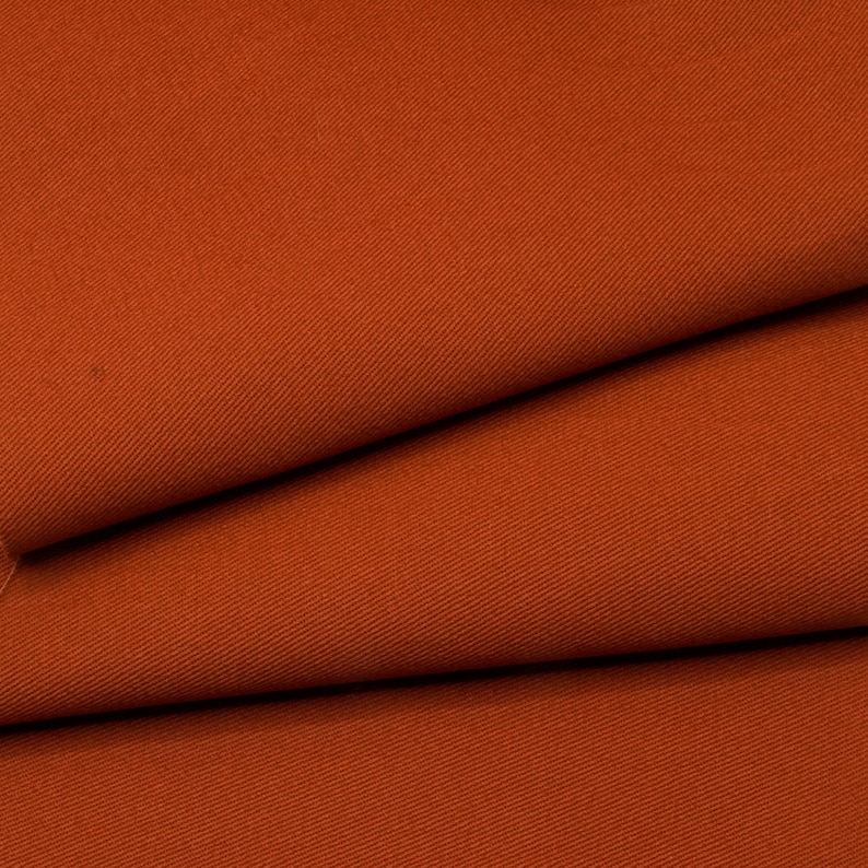 May include: Close-up of a rust-colored fabric with a subtle twill weave. The fabric is folded in thirds, showing the texture and color.