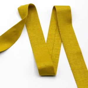 May include: Mustard yellow ribbed knit fabric, possibly elastic, in a wavy pattern against a white background. The fabric appears to be folded and draped, creating a textured visual effect. The color is a warm, earthy tone.