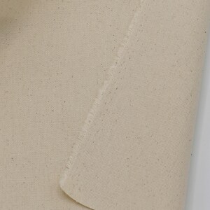 May include: Close-up of a natural beige canvas fabric with a raw edge. The fabric has a subtle texture and a slightly uneven surface.