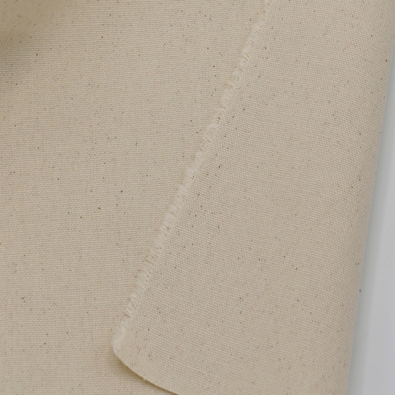 May include: Close-up of a natural beige canvas fabric with a raw edge. The fabric has a subtle texture and a slightly uneven surface.