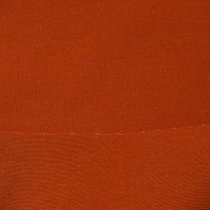 May include: A close-up of a rust-colored fabric with a subtle diagonal weave pattern. The fabric has a smooth, slightly textured surface.