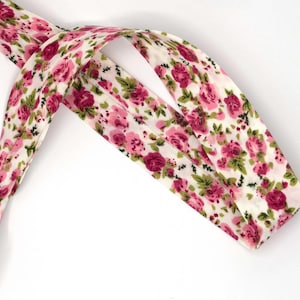 May include: A length of bias tape featuring a repeating floral pattern with pink roses and green leaves on a white background. The fabric strip is folded along the center, showing a consistent print and texture throughout the entire length of the trim.