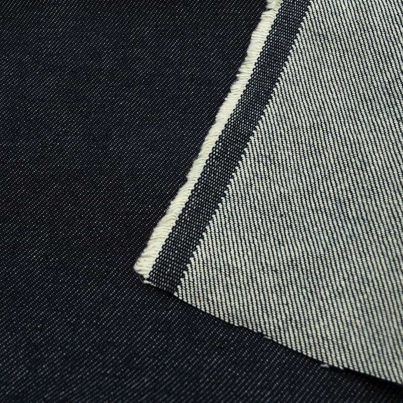 May include: Close-up of dark blue denim fabric with a white selvedge edge. The fabric is woven with a diagonal twill pattern.