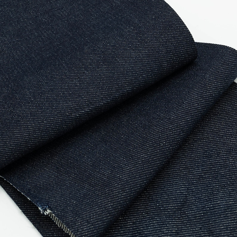 May include: Close-up of dark blue denim fabric with a subtle texture. The fabric is folded in layers, showing the weave and color.