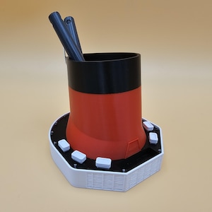 May include: A white and orange 3D printed pen holder with a black top and a hexagonal base. The base has a grid pattern and several small white circles around the edge.