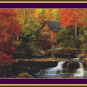 Puede incluir: Un paisaje bordado a punto de cruz representa un molino de madera entre árboles con vibrante follaje otoñal. Una cascada cae en un arroyo, creando una escena serena y colorida. La obra está enmarcada en oro y morado.