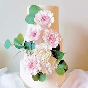 May include: A three-tiered white cake decorated with large pink and white flowers and green leaves. The cake is a classic design, perfect for a wedding or special occasion. The flowers are arranged in a cascading pattern, adding a touch of elegance.
