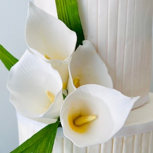 May include: A white wedding cake decorated with white fondant and a cluster of white calla lilies with green leaves.