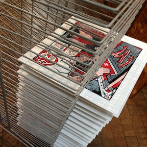 May include: A stack of art prints featuring colorful illustrations, including a luchador mask and a person holding a book. The prints are displayed in a metal wire rack, with visible details in red, white, black, and gray.