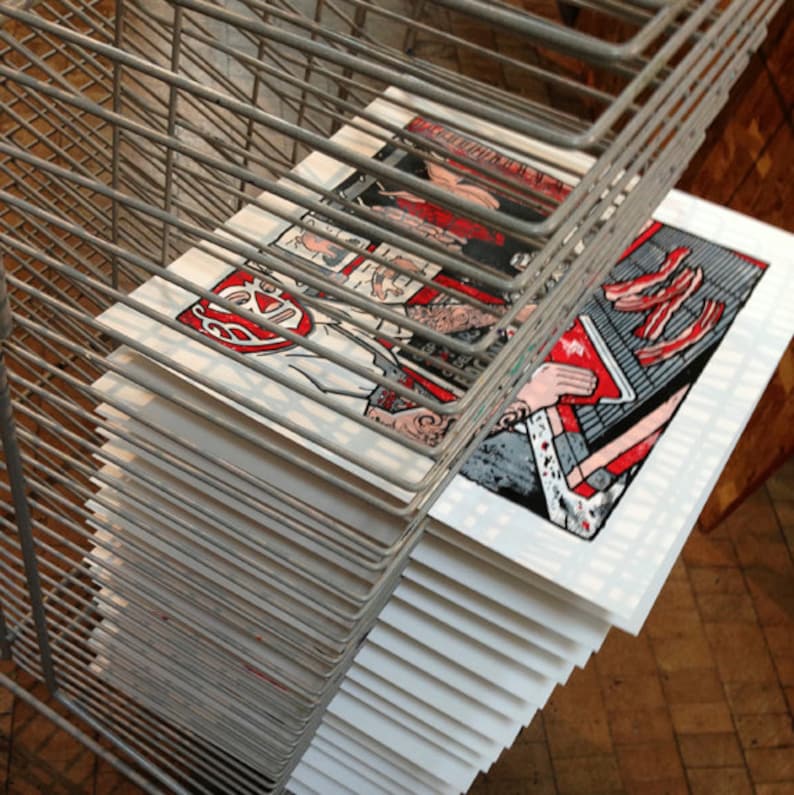 May include: A stack of art prints featuring colorful illustrations, including a luchador mask and a person holding a book. The prints are displayed in a metal wire rack, with visible details in red, white, black, and gray.