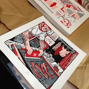 May include: A stack of art prints featuring a colorful illustration of a person in a wrestling mask preparing bacon. The print has red, black, gray, and white elements. Additional prints with similar designs are visible.