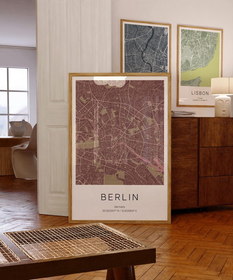 May include: A framed map print of Berlin, Germany, in shades of burgundy and tan. The city map is detailed with streets and parks. The print includes the city name and coordinates. Two other framed city maps are visible in the background.