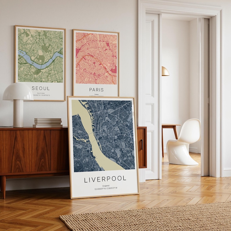 May include: Three framed city map prints. The top two feature maps of Seoul and Paris, with the bottom print showcasing Liverpool. Each map has a different color scheme, with the Liverpool map in blue and beige. The prints are displayed in a room with wooden furniture.