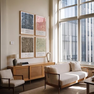 May include: Four framed city map prints hang on a wall. The maps are of Paris, Dallas, Milan, and Frankfurt. The prints have a neutral color palette with a wooden frame. The room also features a wooden cabinet, a sofa, and a chair.