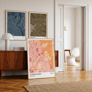May include: Three framed city maps in a room. The maps are in shades of blue, brown, and pink, with the cities of Paris, Krakow, and Copenhagen labeled. The Copenhagen map is leaning against the wall. A wooden cabinet and a white lamp are in the foreground.