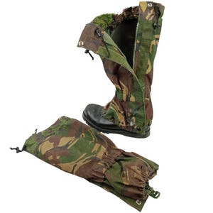 Op de afbeelding: Een paar camouflage-gaiters met een bospatroon in tinten groen, bruin en zwart. Eén gaiter is opengeklapt met een verticale rits en drukknopen, terwijl de andere plat ligt. Beide zijn voorzien van elastische delen en trekkoorden.