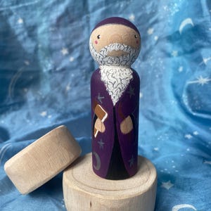 May include: A hand-painted wooden wizard figurine with a purple robe, white beard, and a staff. The wizard stands on a wooden base, with a separate wooden disc nearby. The background is a blue fabric with white stars.
