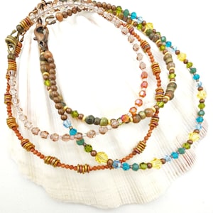 May include: Four beaded necklaces in shades of brown, orange, green, yellow, and blue, displayed on a white seashell. The necklaces feature various bead sizes and textures, with copper-colored clasps and accents. The beads include crystals and small metal rings.