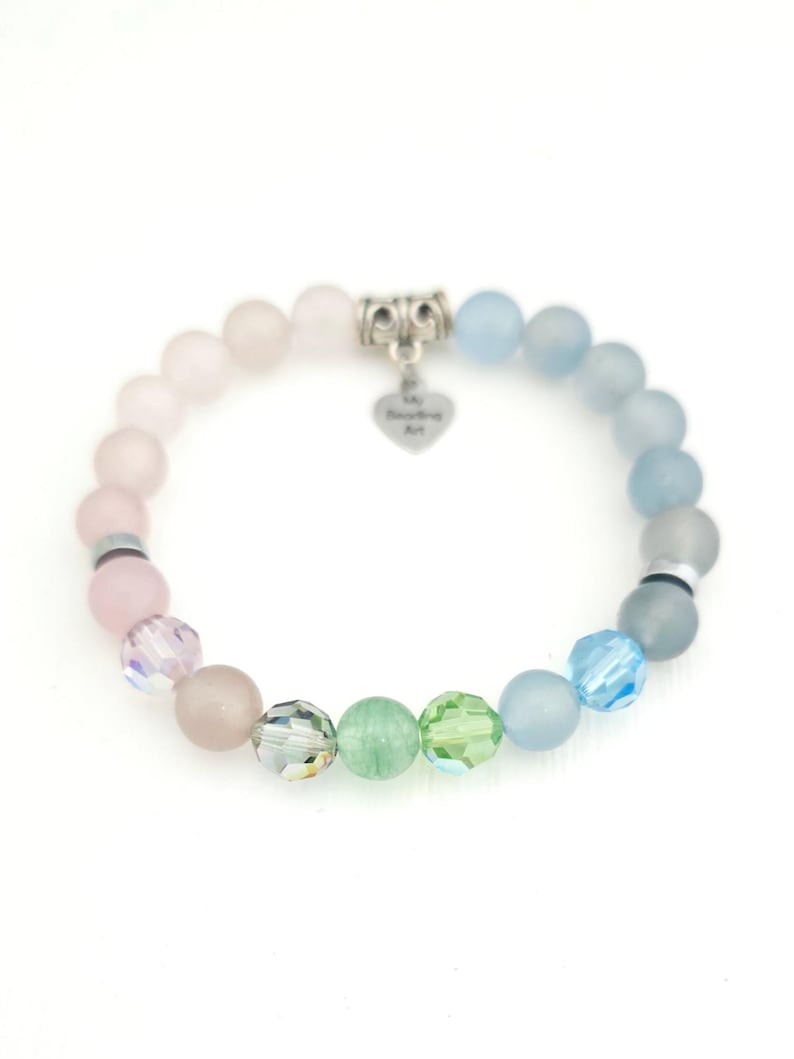 K&ouml;nnte beinhalten: Ein elastisches Armband mit verschiedenen runden Perlen in Rosa-, Blau- und Gr&uuml;nt&ouml;nen. Das Design umfasst matte und facettierte Kristallperlen, silberfarbene Zwischenelemente und einen herzf&ouml;rmigen Anh&auml;nger mit der Gravur My Beading Art.