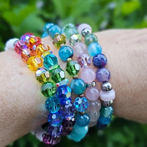 May include: A stack of multiple beaded bracelets featuring a variety of faceted crystal beads in rainbow colors, smooth pastel pink stones, dark blue crackle-textured beads, and metallic silver-toned spacers arranged in a multi-strand design.