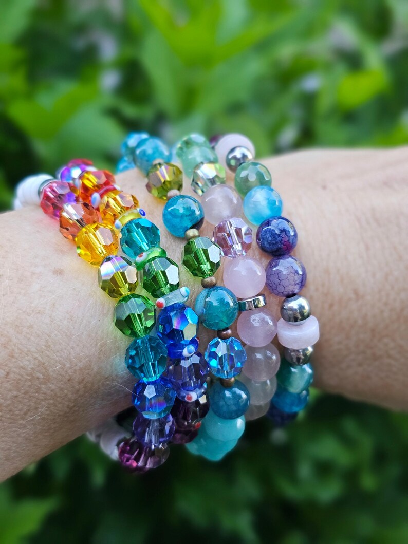 K&ouml;nnte beinhalten: Ein Stapel aus mehreren Perlenarmb&auml;ndern mit einer Vielzahl von facettierten Kristallperlen in Regenbogenfarben, glatten pastellrosa Steinen, dunkelblauen Perlen mit Rissstruktur und metallischen silberfarbenen Zwischenelementen.