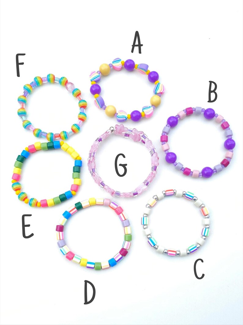 May include: Seven colorful beaded bracelets labeled A through G. Bracelet A has purple and heart beads. B features purple and pink spheres. C has white and striped beads. D uses multicolored cubes. E has bright cubes. F has rainbow beads. G is pink.