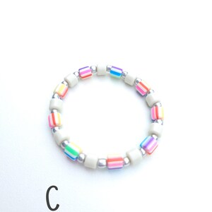 May include: A circular beaded bracelet featuring alternating segments of solid off-white cylindrical beads, metallic silver spherical beads, and multi-colored striped cylindrical beads in shades of pink, orange, yellow, green, blue, and purple. The letter C is visible.